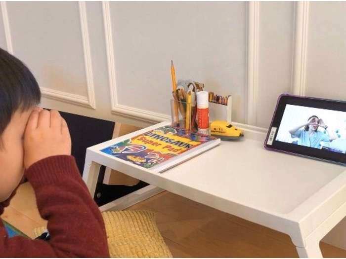 A pre-school boy sits in his room and plays along with his teacher during online lessons.