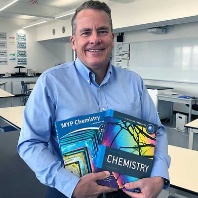 The Chemistry teacher is standing in his lab holding two textbooks he authored.