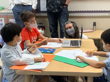 Students and teachers gather around a laptop for an online learning session.