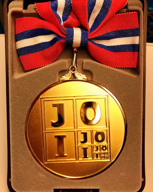 A round, gold medal with a red, white, and blue ribbon sitting in a brown velvet box. The medal reads JOI