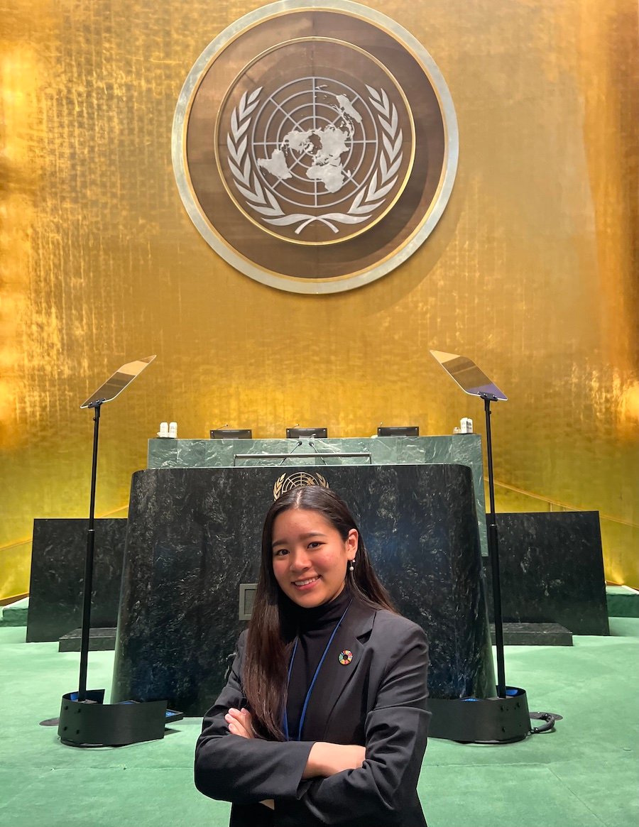 A young woman stands in front of a podium and seal of the United Nations that is hung on a golden wall.