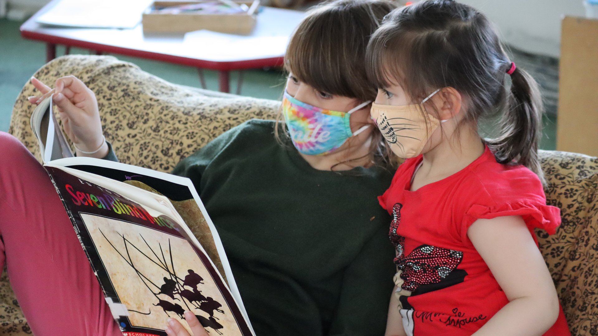 An older child in a green sweatshirt and tie-dye mask reads a picture book to a younger child in a red T-shirt and tiger mask as they sit on a small sofa
