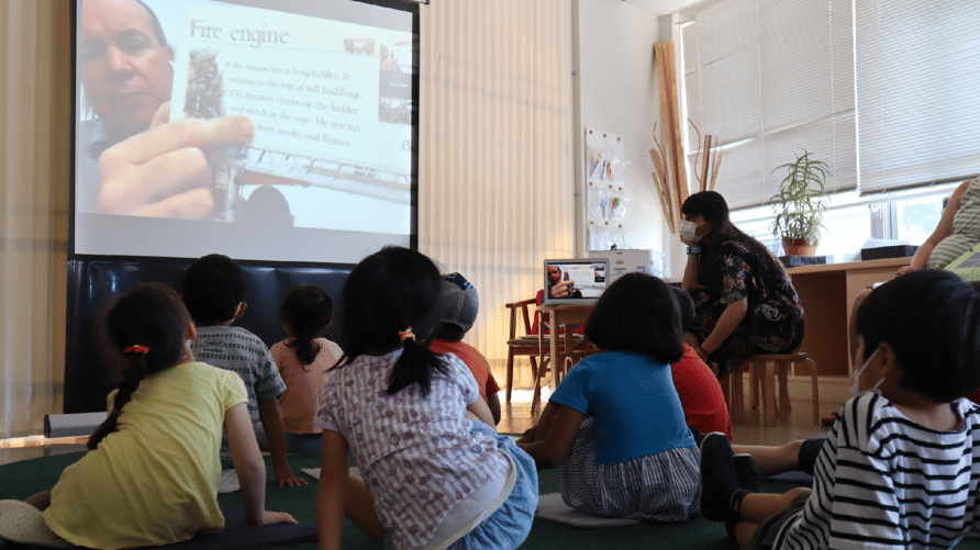 Primary students sit on the floor and gaze at an instructor on a big screen.