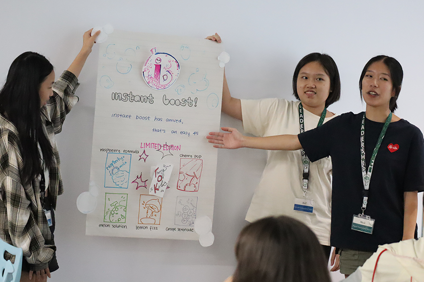 Three girls present their idea for the leadership conference using a poster with colorful graphics.