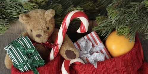 An orange, teddy bear, candy cane and small presents inside a Christmas stocking.